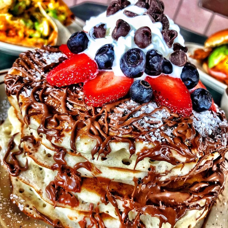 Stack of pancakes with chocolate drizzle, powdered sugar, whipped cream, strawberries, blueberries, and chocolate chips.
