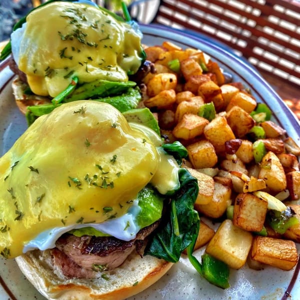 Two Eggs Benedict with hollandaise sauce, spinach, avocado, and bacon, served with home fries.