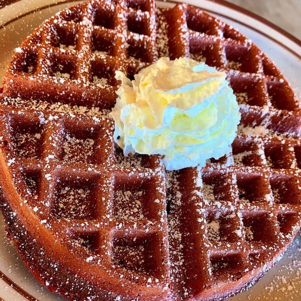 A red velvet waffle dusted with powdered sugar and topped with whipped cream on a plate.