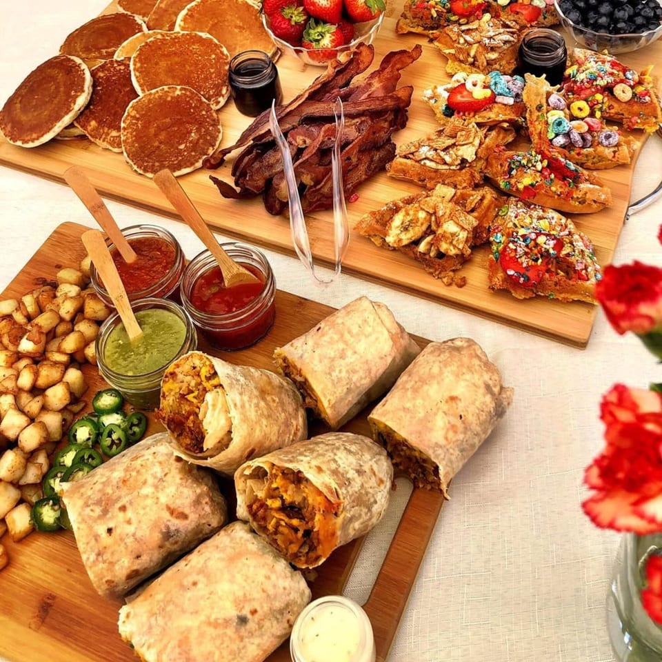 A breakfast spread with pancakes, bacon, waffles, breakfast burritos, home fries, and sauces on wooden boards.