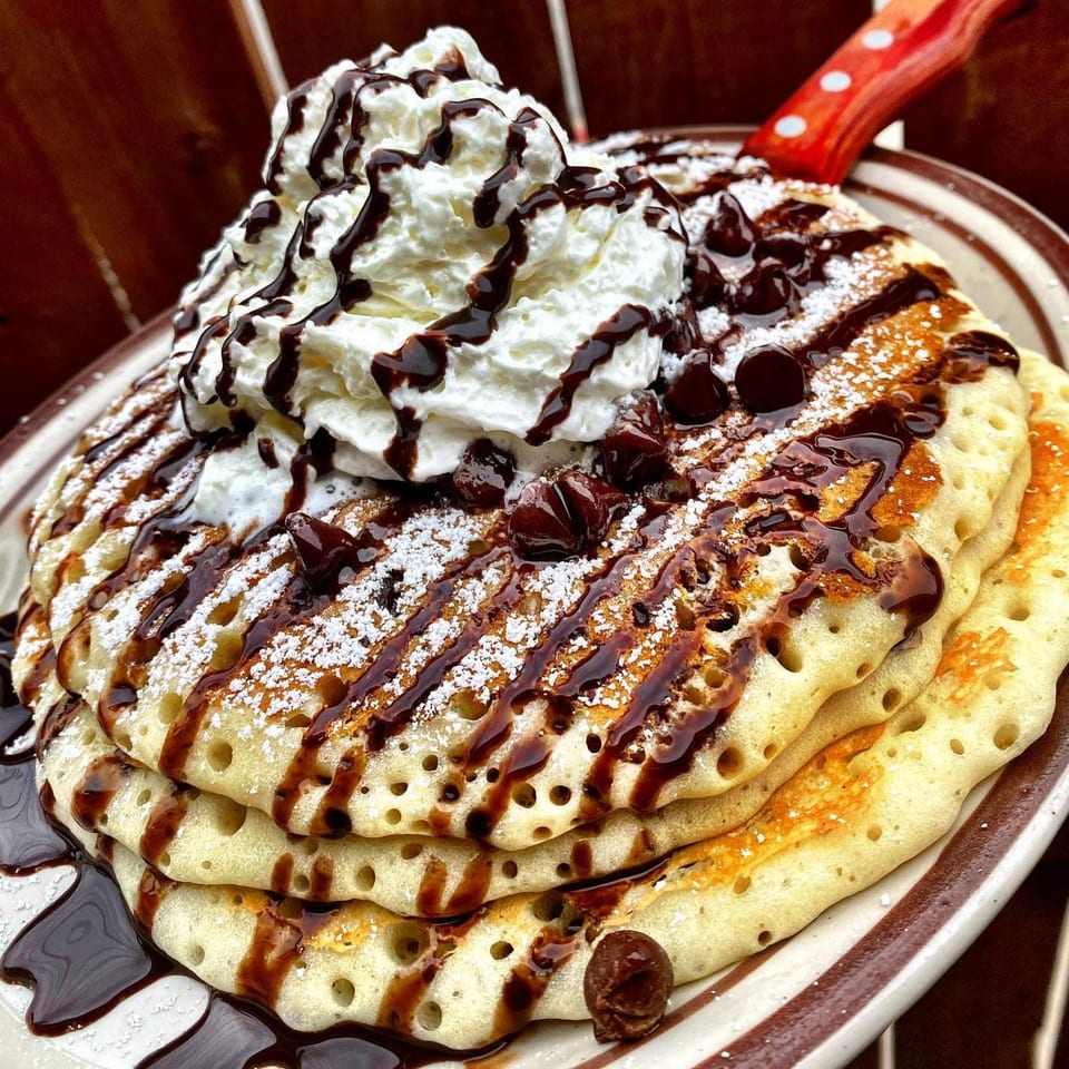 Stack of pancakes topped with whipped cream, chocolate syrup, chocolate chips, and powdered sugar.