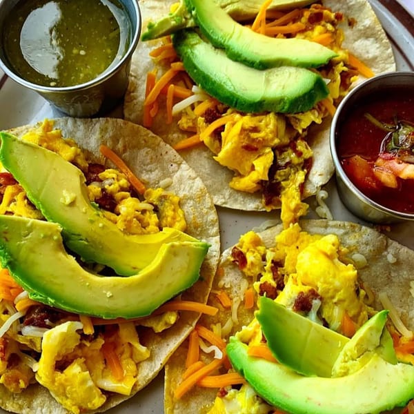 Three breakfast tacos with scrambled eggs, cheese, avocado slices, and two dipping sauces.